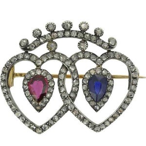 May include: A silver and gold brooch with two intertwined heart shapes set with diamonds. The hearts are accented with a ruby and a sapphire.