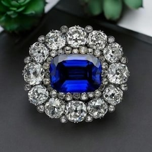 May include: A large, oval-shaped brooch with a large, rectangular, dark blue sapphire surrounded by a halo of smaller, round, white diamonds.