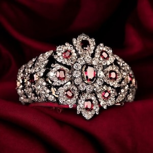 Op de afbeelding: Een zilveren armband met diamanten en een groot rood robijn in het midden. De armband is versierd met kleinere robijnen en diamanten, wat resulteert in een luxueus en elegant ontwerp.