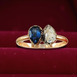 Puede incluir: Un anillo de oro con dos piedras preciosas en forma de pera. Una es azul oscuro, la otra es transparente. El anillo se presenta sobre un fondo rojo, resaltando el contraste de las piedras y el oro.