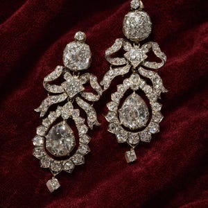 以下が含まれることがあります： 装飾的なダイヤモンドのイヤリングのペア。各イヤリングは、下部に大きな洋梨型のダイヤモンドがあり、小さな石で囲まれています。ダイヤモンドが付いた弓型のデザインが、上部の正方形にカットされたダイヤモンドに接続されています。濃い赤色のベルベットの背景。