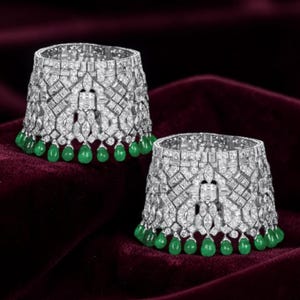 May include: Two silver-toned cuff bracelets encrusted with sparkling clear stones. Each bracelet features a geometric design and is adorned with a row of green teardrop-shaped gemstones. The bracelets are displayed on a deep red velvet surface.