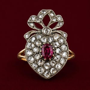 May include: A heart-shaped ring with a central ruby surrounded by numerous clear gemstones. The ring features a bow-shaped design at the top and a gold band. The ring is set against a deep red background.