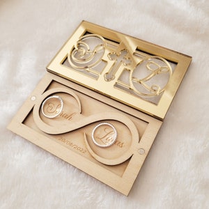 Peut inclure: Une boîte à alliances en bois avec un motif en forme d'infini. La boîte est ouverte et deux alliances sont à l'intérieur. La boîte est gravée des noms "Sarah" et "Lucas" et de la date "20/08/2022". Le dessus de la boîte est un miroir doré avec une croix et un motif décoratif.