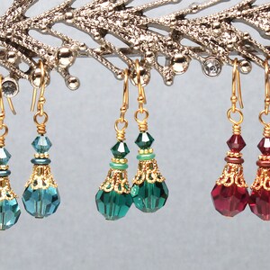 May include: Three pairs of dangle earrings with gold-colored hooks. Each earring features a faceted teardrop-shaped crystal in teal, emerald green, and ruby red, accented with gold filigree and smaller crystals.