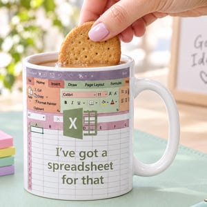 Könnte beinhalten: Ein weißer Keramikbecher mit einem aufgedruckten Microsoft Excel-Tabellen-Design, dem Text 'I've got a spreadsheet for that' und dem Excel-Logo. Der Becher ist mit einem braunen Getränk gefüllt, in das ein runder Keks eingetaucht wird.