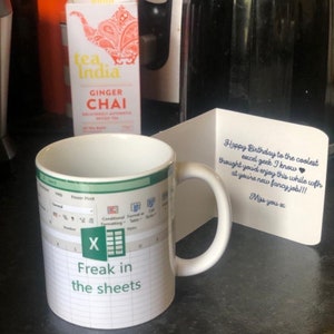 K&ouml;nnte beinhalten: Eine wei&szlig;e Tasse mit einem gr&uuml;nen und wei&szlig;en Excel-Tabellenblatt-Design. Die Tasse tr&auml;gt den Text "Freak in the sheets". Im Hintergrund befindet sich eine Geburtstagskarte mit blauer Schrift.