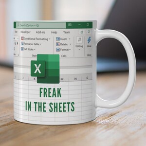 K&ouml;nnte beinhalten: Eine wei&szlig;e Kaffeetasse mit einem gr&uuml;nen Excel-Tabellenblatt-Design. Die Tasse tr&auml;gt den Text "FREAK IN THE SHEETS".