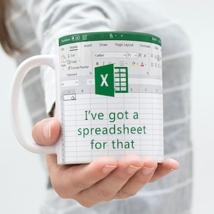 Może przedstawiać: Biały ceramiczny kubek z nadrukiem graficznym interfejsu arkusza kalkulacyjnego Microsoft Excel. Projekt zawiera zielone logo Excel oraz tekst "I've got a spreadsheet for that" w ciemnozielonej czcionce wyśrodkowany na powierzchni kubka.