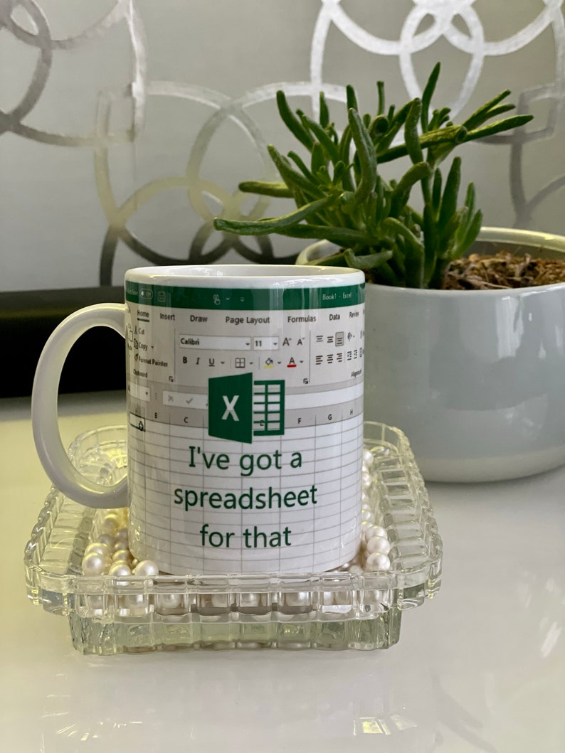 K&ouml;nnte beinhalten: Eine wei&szlig;e Kaffeetasse mit einem gr&uuml;nen Microsoft Excel-Tabellenkalkulationssymbol und dem Text "Ich habe eine Tabelle daf&uuml;r". Die Tasse steht auf einem durchsichtigen Glasuntersetzer mit wei&szlig;en Perlen.