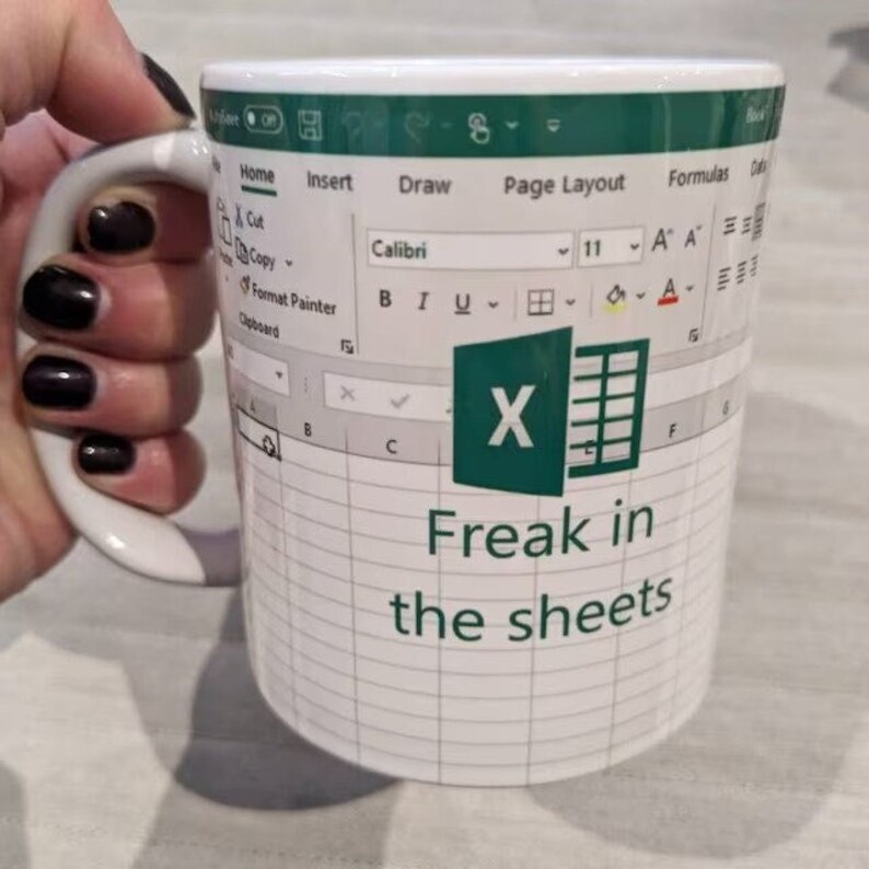 K&ouml;nnte beinhalten: Eine wei&szlig;e Keramiktasse mit einem gr&uuml;nen Microsoft Excel-Logo und einem Tabellenblatt-Design. Die Tasse tr&auml;gt den Text "Freak in the sheets".