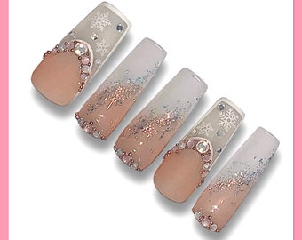 Op de afbeelding: Vijf lange, witte, ombre press-on nagels met een glitterontwerp en strass. De nagels zijn versierd met sneeuwvlokken en hebben een verloop van wit naar lichtroze.
