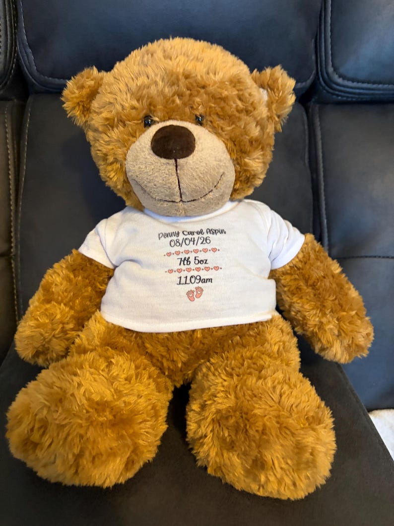 Puede incluir: Un oso de peluche marr&oacute;n que lleva una camiseta blanca. La camiseta presenta el texto Penny Carol Aspin, la fecha 08/04/26, el peso 7lb 5oz, la hora 11:09am y un peque&ntilde;o gr&aacute;fico de huellas de beb&eacute;, todo impreso en tinta negra y roja.
