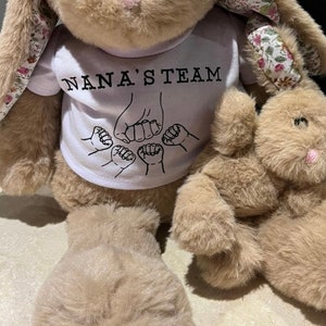 Puede incluir: Un conejo de peluche beige grande con orejas forradas de flores lleva una camiseta blanca con el texto NANA'S TEAM y un gr&aacute;fico de cinco pu&ntilde;os chocando, junto a un conejo de peluche beige m&aacute;s peque&ntilde;o con nariz rosa.