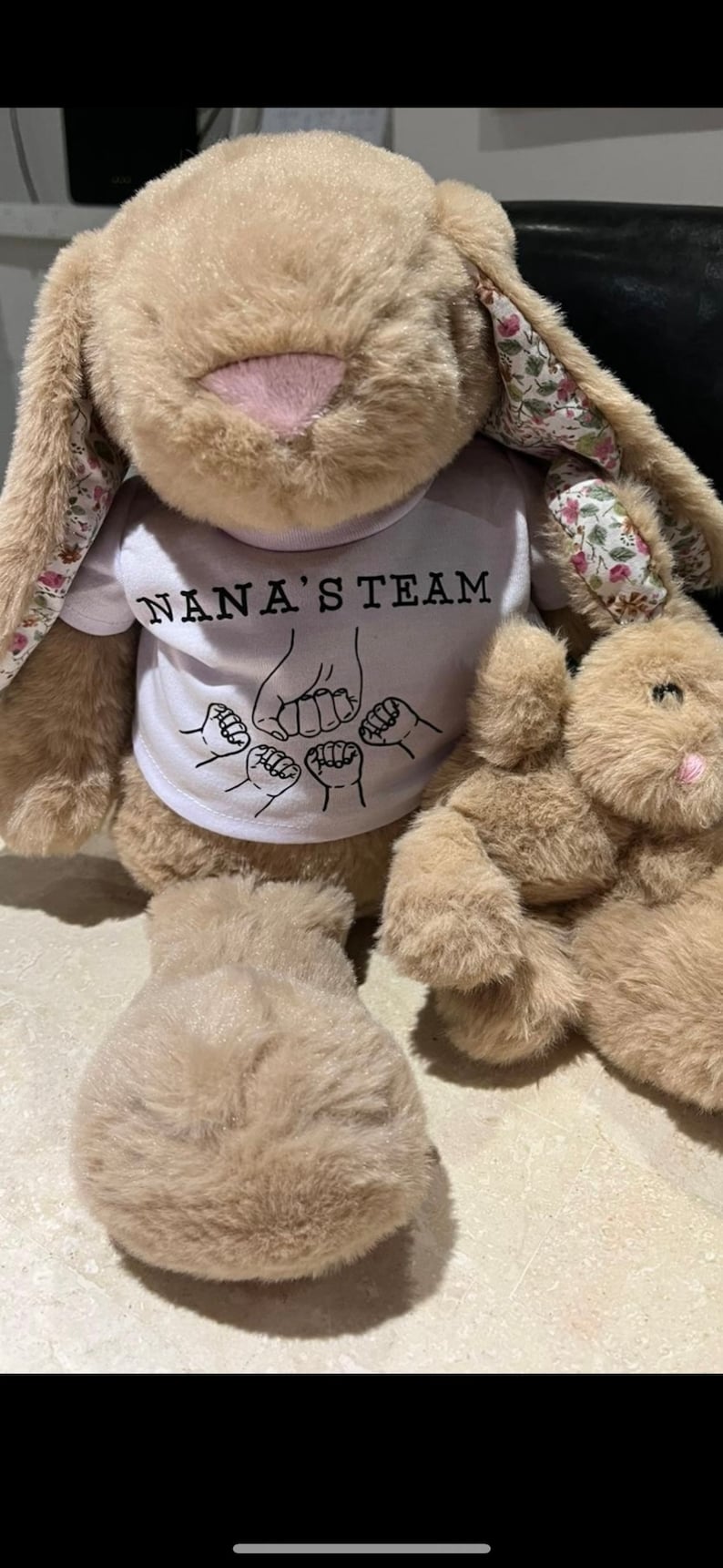 Puede incluir: Un conejo de peluche beige grande con orejas forradas de flores lleva una camiseta blanca con el texto NANA'S TEAM y un gr&aacute;fico de cinco pu&ntilde;os chocando, junto a un conejo de peluche beige m&aacute;s peque&ntilde;o con nariz rosa.