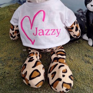 Puede incluir: Un peluche con estampado de leopardo que lleva una camiseta blanca de manga corta. La camiseta presenta un contorno de coraz&oacute;n rosa y el texto Jazzy impreso en rosa en la parte delantera. El juguete tiene pelaje suave beige con manchas negras y marrones.