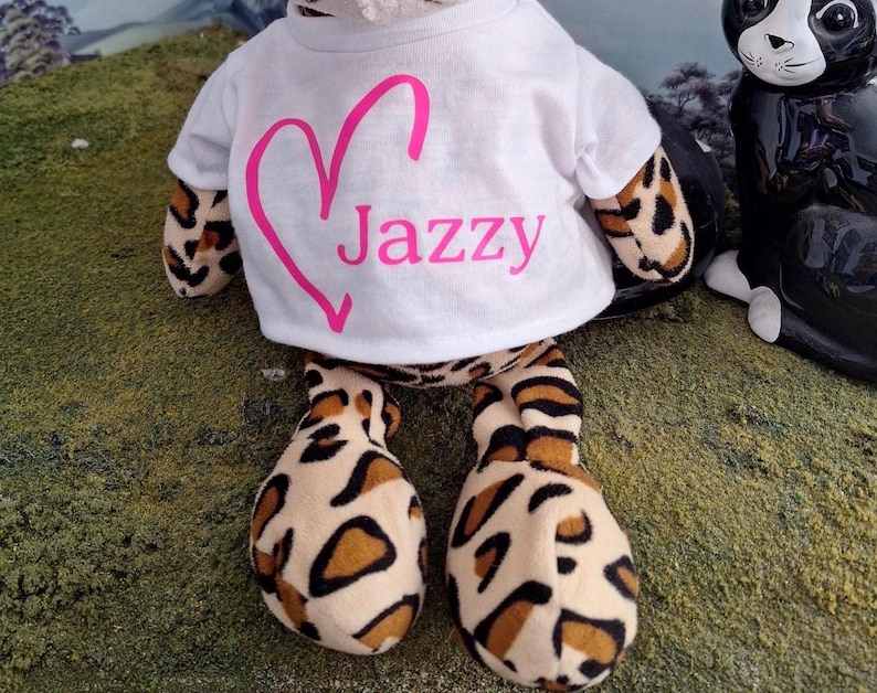 Puede incluir: Un peluche con estampado de leopardo que lleva una camiseta blanca de manga corta. La camiseta presenta un contorno de coraz&oacute;n rosa y el texto Jazzy impreso en rosa en la parte delantera. El juguete tiene pelaje suave beige con manchas negras y marrones.