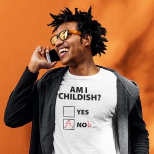 May include: White t-shirt with the text "AM I CHILDISH?" and a check box next to "YES" and a penis graphic next to "NO". The person in the image is wearing sunglasses and a black jacket.