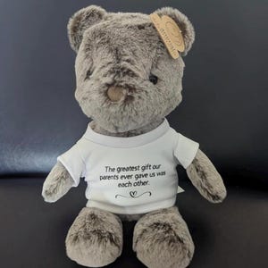 Puede incluir: Un oso de peluche de suave pelaje gris que lleva una camiseta blanca. La camiseta tiene el texto impreso en negro: The greatest gift our parents ever gave us was each other, con un peque&ntilde;o s&iacute;mbolo de coraz&oacute;n debajo. Una etiqueta beige est&aacute; sujeta a la oreja.