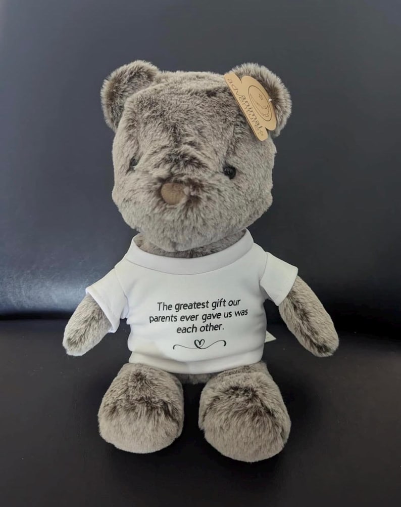 Puede incluir: Un oso de peluche de suave pelaje gris que lleva una camiseta blanca. La camiseta tiene el texto impreso en negro: The greatest gift our parents ever gave us was each other, con un peque&ntilde;o s&iacute;mbolo de coraz&oacute;n debajo. Una etiqueta beige est&aacute; sujeta a la oreja.
