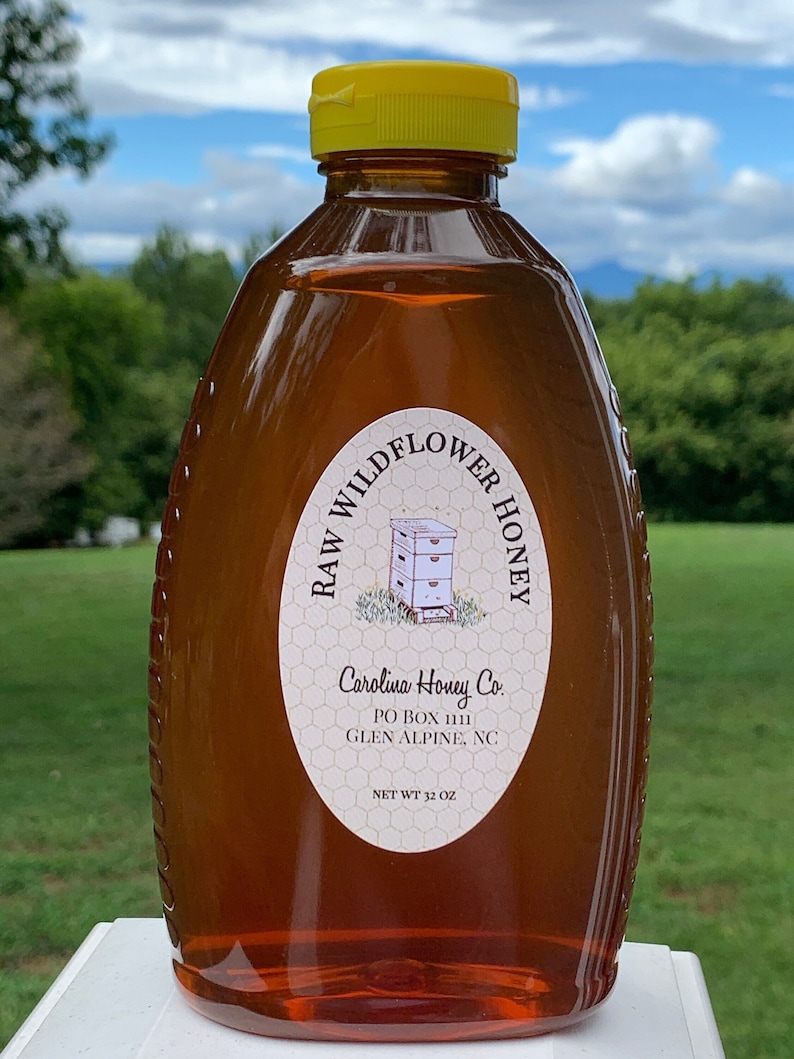 May include: A clear glass bottle of raw wildflower honey with a yellow lid. The label reads "RAW WILDFLOWER HONEY" and "Carolina Honey Co. PO BOX 1111 GLEN ALPINE, NC NET WT 32 OZ".