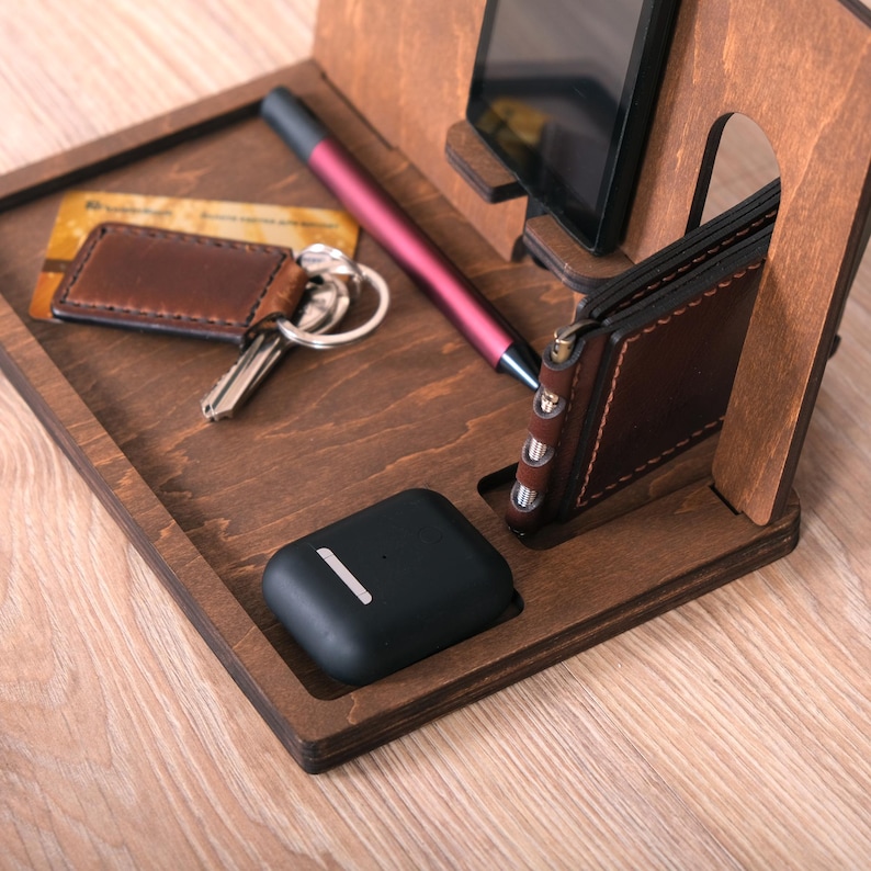 Pu&ograve; includere: Un supporto per telefono in legno con slot per telefono, portafoglio, chiavi e penna. Il supporto &egrave; in legno marrone scuro e ha una finitura naturale. C'&egrave; una custodia per telefono nera, un portafoglio in pelle marrone, un set di chiavi e una penna rosa sul supporto.