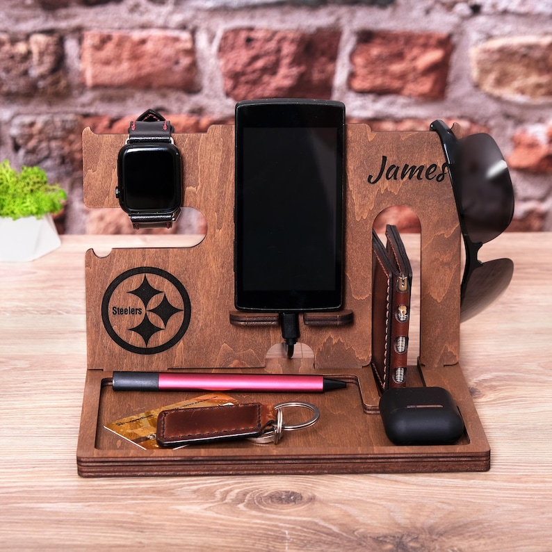 Pu&ograve; includere: Un supporto per telefono in legno con un nome personalizzato "James" e il logo dei Pittsburgh Steelers. Ha slot per un telefono, un orologio, occhiali da sole e un portafoglio.