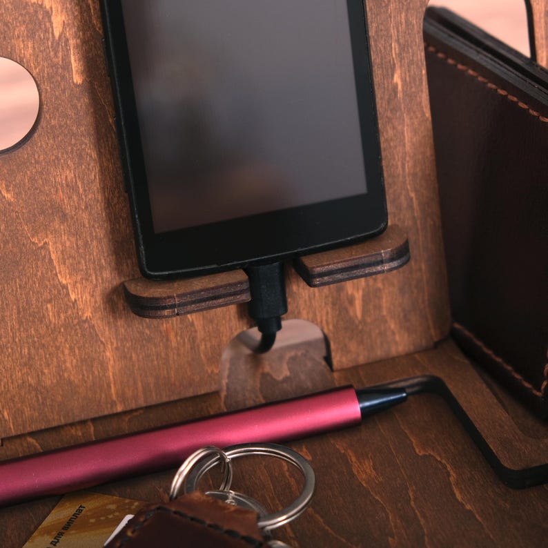 Pu&ograve; includere: Un supporto per telefono in legno con un telefono, una penna, chiavi e un portafoglio. Il telefono &egrave; in carica con un cavo nero.