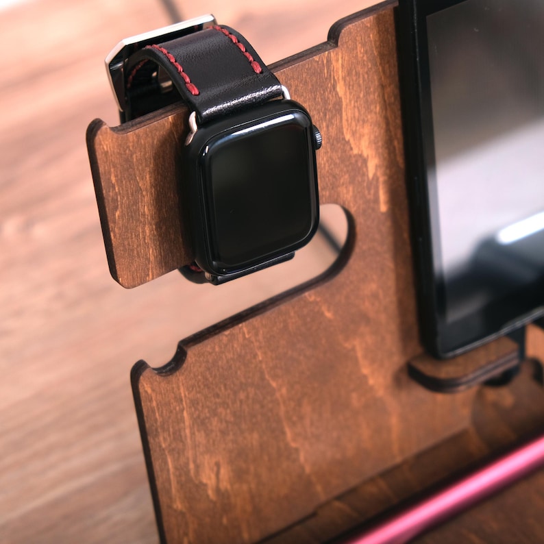 Pu&ograve; includere: Un supporto in legno con una fessura per un orologio e un telefono. L'orologio ha un cinturino in pelle nera con cuciture rosse. Il telefono &egrave; nero e ha uno schermo.
