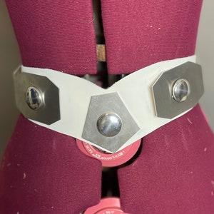 Puede incluir: Un cinturón blanco con detalles metálicos plateados. El cinturón presenta placas metálicas geométricas con remaches plateados redondeados. El cinturón se muestra en un maniquí burdeos.