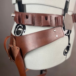 May include: A brown leather belt with a silver buckle and multiple pouches. The belt is worn on a mannequin and has a holster attached to the side.