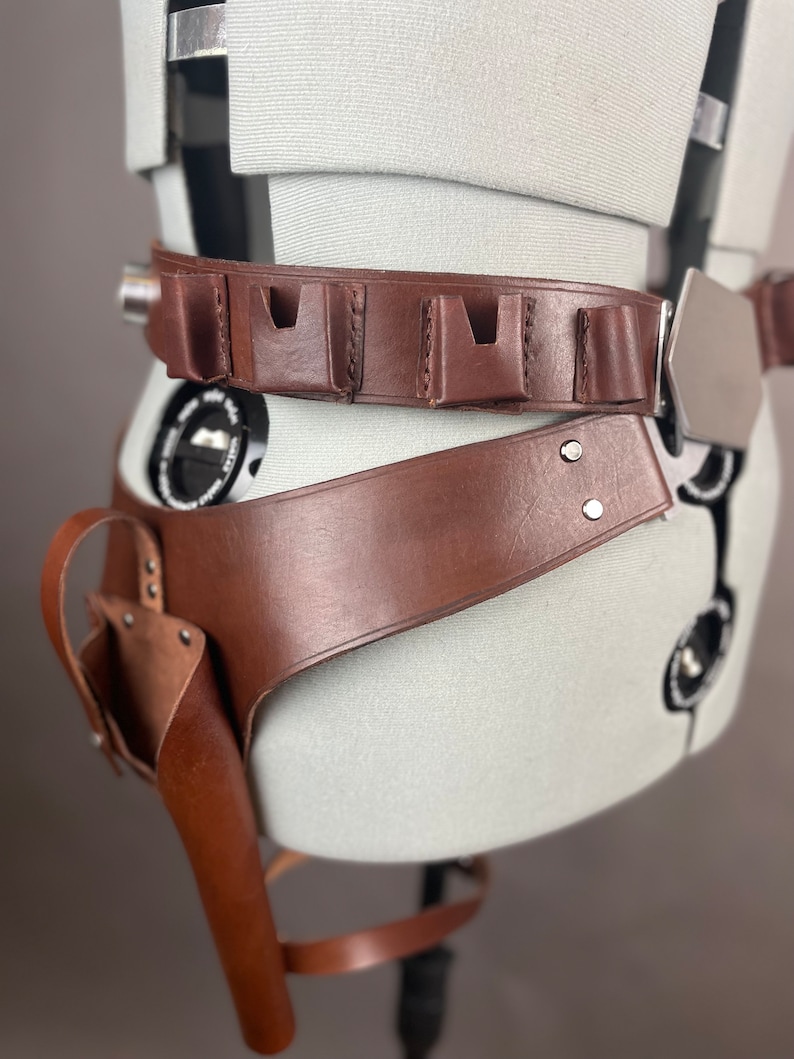 May include: A brown leather belt with a silver buckle and multiple pouches. The belt is worn on a mannequin and has a holster attached to the side.