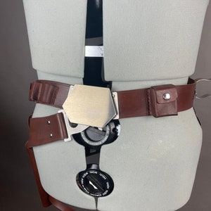 May include: A brown leather belt with a silver buckle and a small brown leather pouch with a metal button. The belt is worn on a white mannequin.