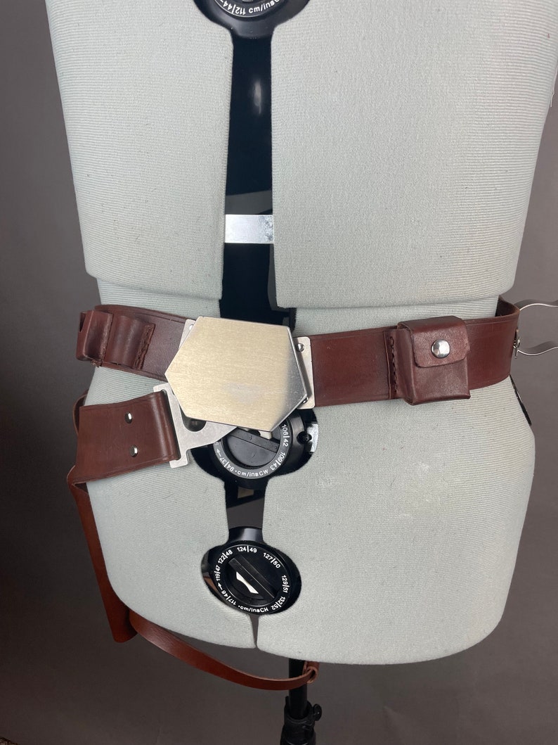 May include: A brown leather belt with a silver buckle and a small brown leather pouch with a metal button. The belt is worn on a white mannequin.