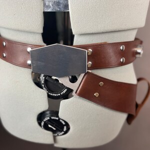 May include: A brown leather belt with a silver buckle and a metal plate. The belt has a silver buckle with a small dial and a metal plate with a smooth surface. The belt is worn on a mannequin.