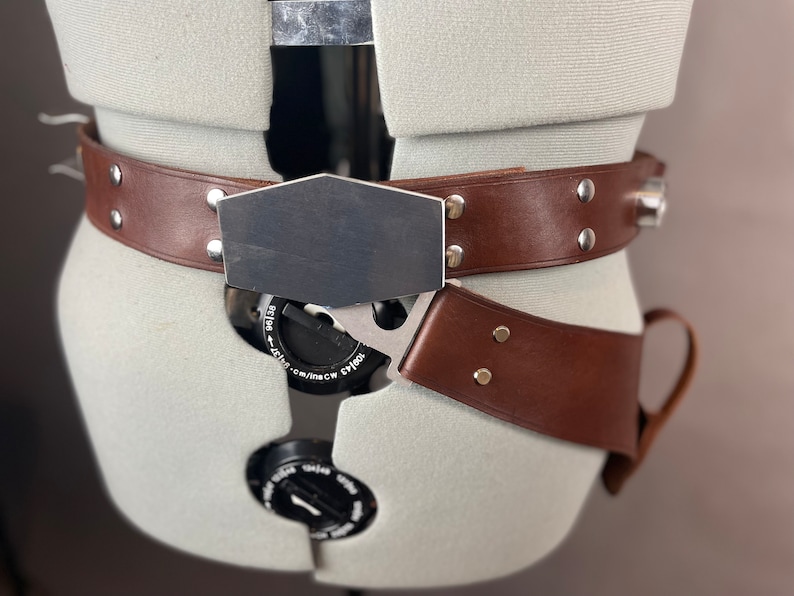 May include: A brown leather belt with a silver buckle and a metal plate. The belt has a silver buckle with a small dial and a metal plate with a smooth surface. The belt is worn on a mannequin.
