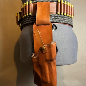 Puede incluir: Un cintur&oacute;n de cuero marr&oacute;n con cartuchos adjuntos y una funda a juego. El cintur&oacute;n est&aacute; asegurado con un material similar a una cuerda negra y gris. La funda est&aacute; sujeta al cintur&oacute;n con una correa de cuero y un bot&oacute;n de lat&oacute;n.