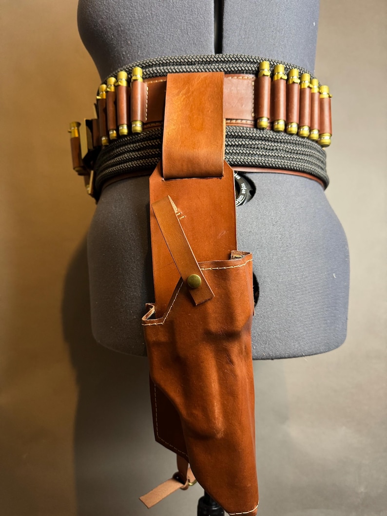 Puede incluir: Un cintur&oacute;n de cuero marr&oacute;n con cartuchos adjuntos y una funda a juego. El cintur&oacute;n est&aacute; asegurado con un material similar a una cuerda negra y gris. La funda est&aacute; sujeta al cintur&oacute;n con una correa de cuero y un bot&oacute;n de lat&oacute;n.