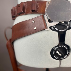 May include: A brown leather belt with a metal buckle and a holster attached. The belt has multiple loops and a metal plate with markings in inches and centimeters.