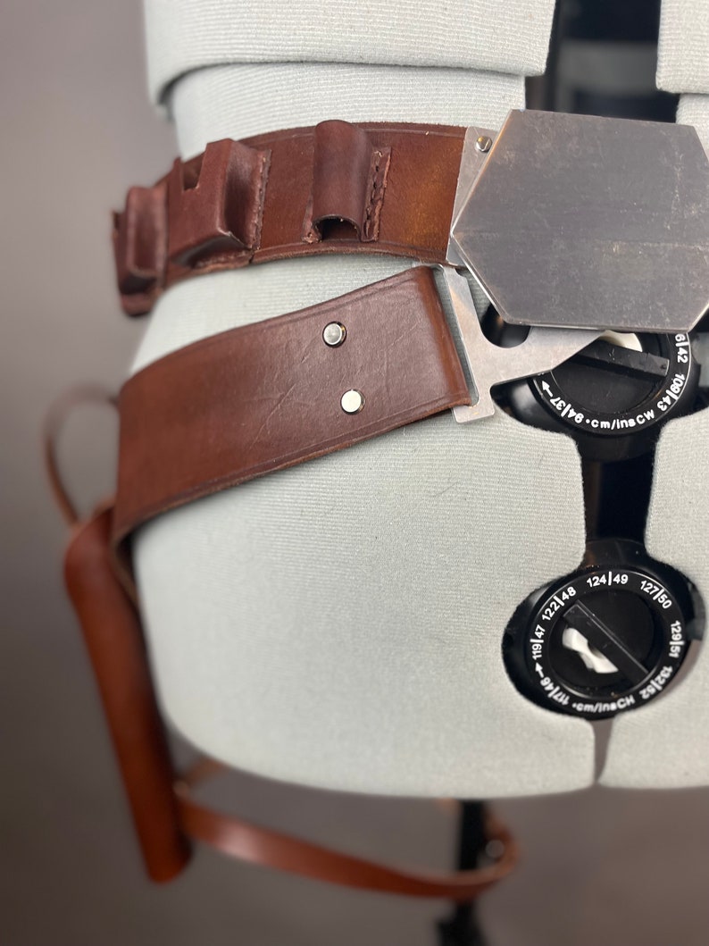 May include: A brown leather belt with a metal buckle and a holster attached. The belt has multiple loops and a metal plate with markings in inches and centimeters.