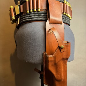 Puede incluir: Un cintur&oacute;n de cuero marr&oacute;n con una funda y portacartuchos. El cintur&oacute;n presenta un dise&ntilde;o de cuerda negra y gris y detalles dorados. La funda est&aacute; dise&ntilde;ada para sujetar una pistola. El cintur&oacute;n se exhibe en un maniqu&iacute;.