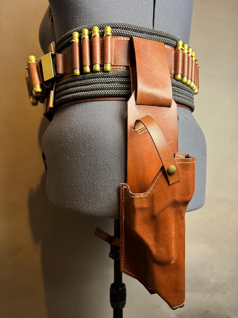 Puede incluir: Un cintur&oacute;n de cuero marr&oacute;n con una funda y portacartuchos. El cintur&oacute;n presenta un dise&ntilde;o de cuerda negra y gris y detalles dorados. La funda est&aacute; dise&ntilde;ada para sujetar una pistola. El cintur&oacute;n se exhibe en un maniqu&iacute;.