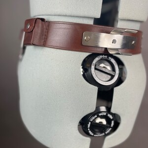 May include: A brown leather belt with a silver buckle and metal accents. The belt is on a white mannequin.