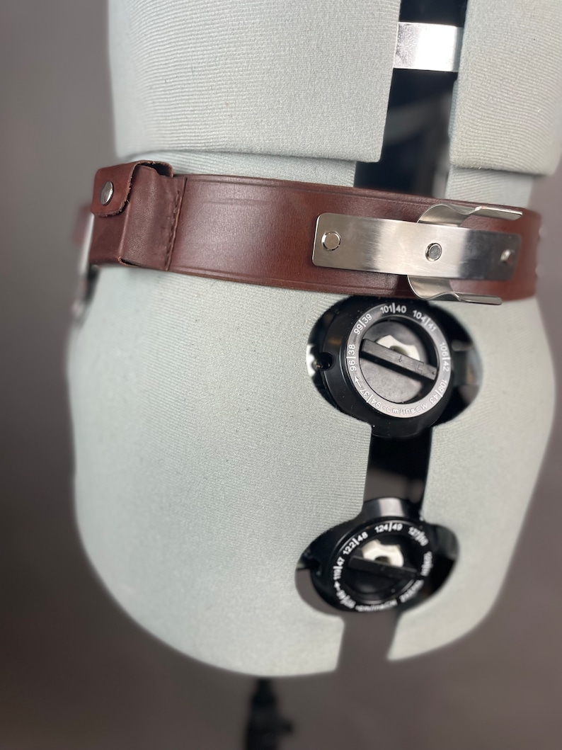 May include: A brown leather belt with a silver buckle and metal accents. The belt is on a white mannequin.