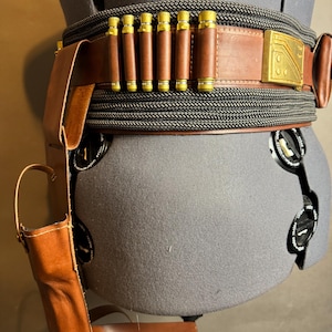 Puede incluir: Un cintur&oacute;n de cuero marr&oacute;n con porta cartuchos y una funda adjuntos. El cintur&oacute;n presenta una hebilla dorada y est&aacute; envuelto con un material similar a una cuerda negra y gris. La funda tambi&eacute;n es de cuero marr&oacute;n.