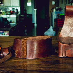 Puede incluir: Tres cinturones de cuero marrón, uno con un diseño tejido, otro con una hebilla y el último con un lazo. Los cinturones están sobre una superficie de madera.