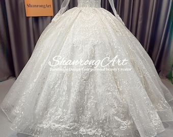 Puede incluir: Un vestido de novia blanco de manga larga, adornado con detalles de cuentas y bordados. El vestido presenta un cuello alto y una falda amplia de estilo de baile. El corpiño está adornado y el vestido se exhibe en un maniquí.