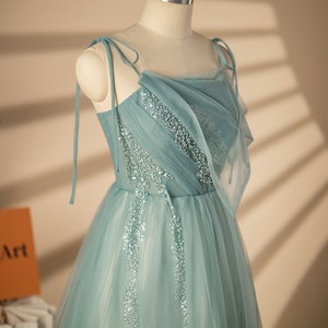 May include: A light blue tulle dress with a draped neckline and beaded embellishments. The dress has thin straps that tie at the shoulders. The dress is on a white mannequin. The text "ShanrongArt Handmade Design Your personal beauty Creator" is at the bottom of the image.