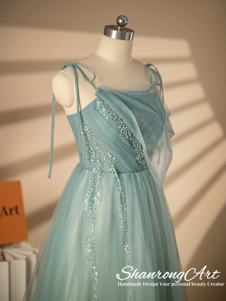 May include: A light blue tulle dress with a draped neckline and beaded embellishments. The dress has thin straps that tie at the shoulders. The dress is on a white mannequin. The text "ShanrongArt Handmade Design Your personal beauty Creator" is at the bottom of the image.