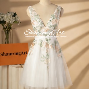 Puede incluir: Vestido blanco hasta la rodilla con cuello en V y bordado floral. El vestido presenta una falda de tul y se exhibe en un maniquí. El texto "ShanrongArt" es visible en el vestido y en una caja.